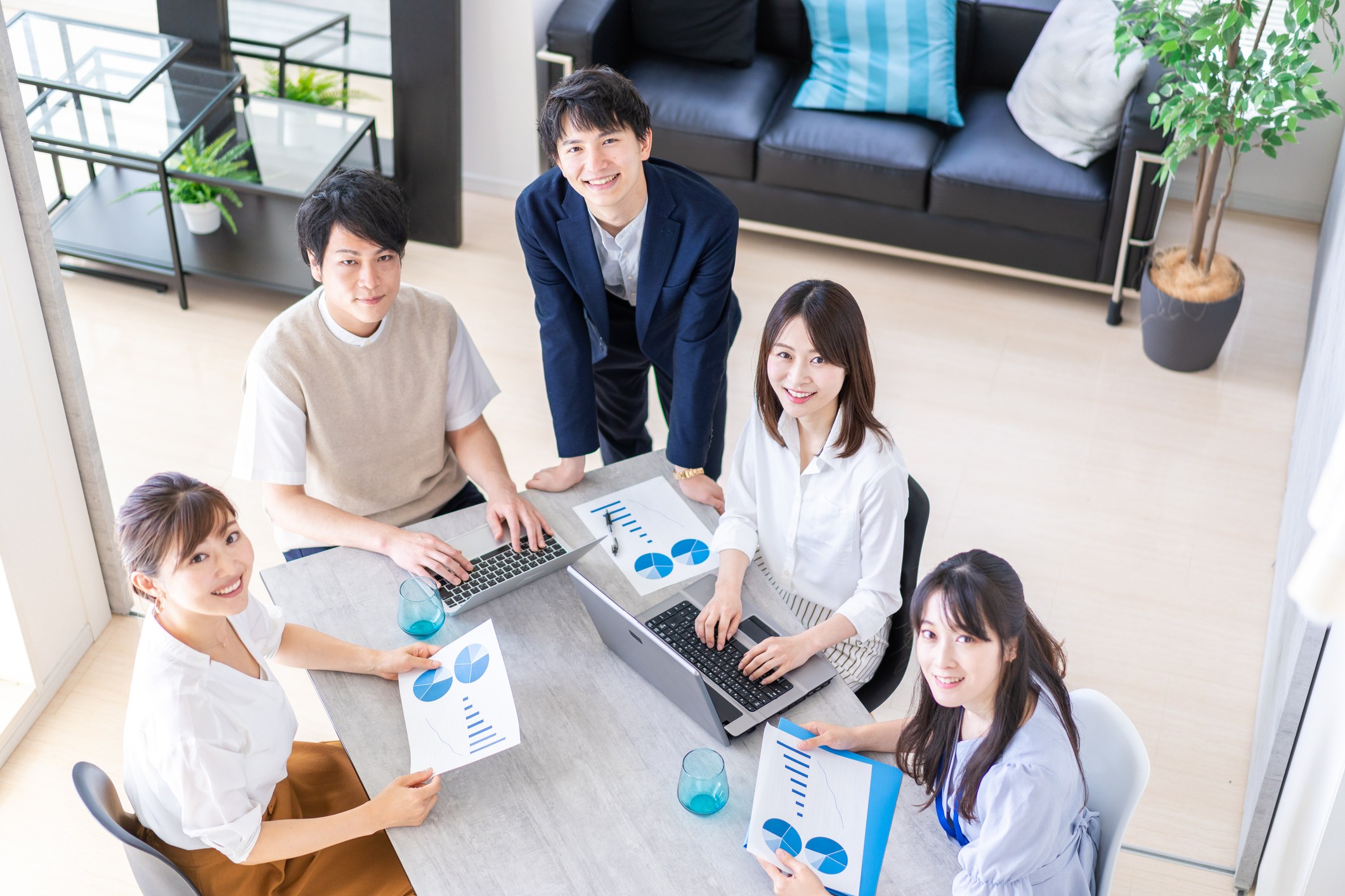 明るいオフィスのテーブルを囲み、資料とノートPCを手にミーティングをする5人のビジネスパーソン