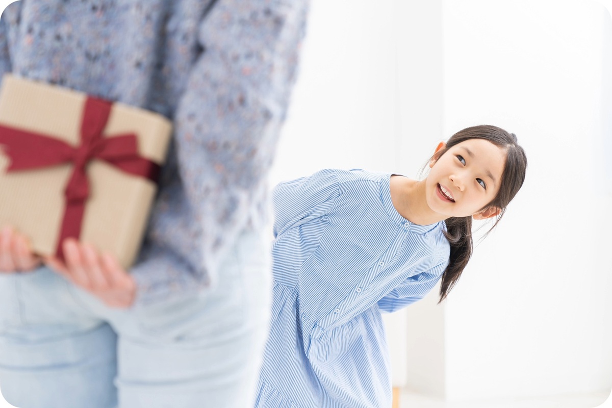 プレゼントを背中に隠した大人の後ろで、嬉しそうに振り返る女の子